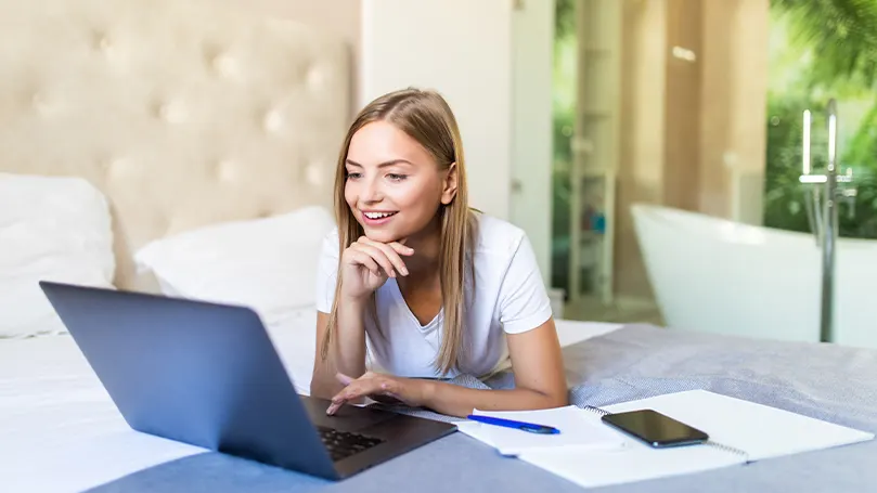 Et billede af en kvinde, der ligger på sin seng og arbejder på sin laptop.